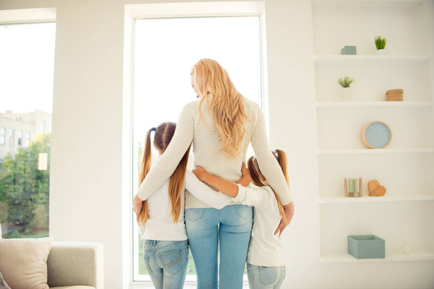 Rear back behind view of nice adorable stylish trendy slim people mom mommy mum girls hugging in front of window in light white interior room hotel indoors - Photo, Image