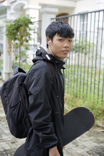 junger positiver Teenager mit Skateboard, der Musik im Kopfhörer hört - Foto, Bild