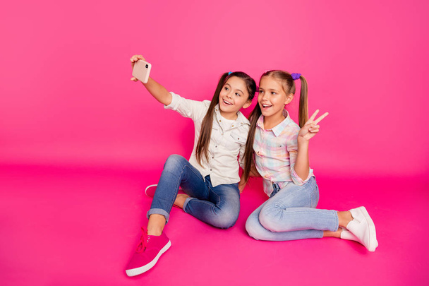 Close up photo two people little age she her girls hold hands arms telephones make take selfies v-sign sit floor  wear casual jeans denim checkered plaid shirts isolated rose vibrant background - 写真・画像