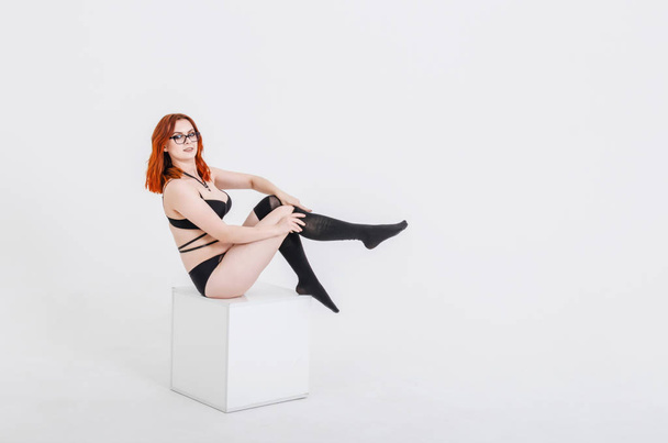 Femme sexy aux cheveux roux en lingerie sombre et bas assis sur cube blanc. Studio, sexualité, séduction
 - Photo, image