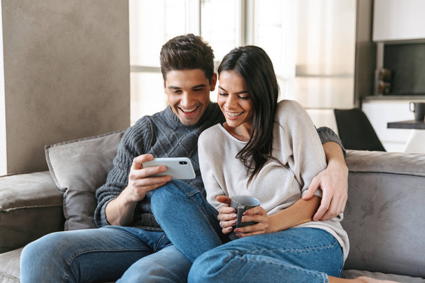 Happy young couple sitting on a couch at home, drinking tea, looking at mobile phone - Фото, изображение
