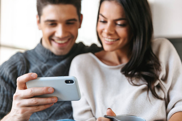 Happy young couple sitting on a couch at home, drinking tea, looking at mobile phone - Foto, imagen