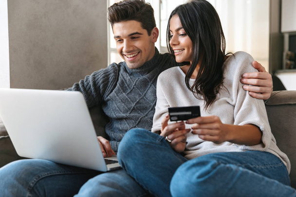 Happy young couple sitting on a couch at home, using laptop computer, showing credit card - Photo, image
