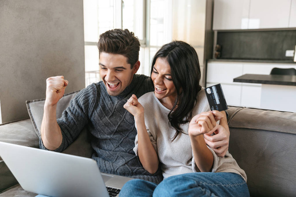 Happy young couple sitting on a couch at home, using laptop computer, showing credit card - Foto, imagen