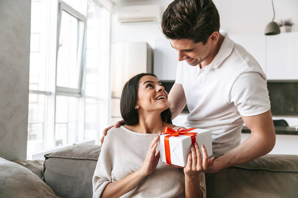 Happy lovely young couple celebrating together while sitting on a couch at home, giving presents - Foto, imagen