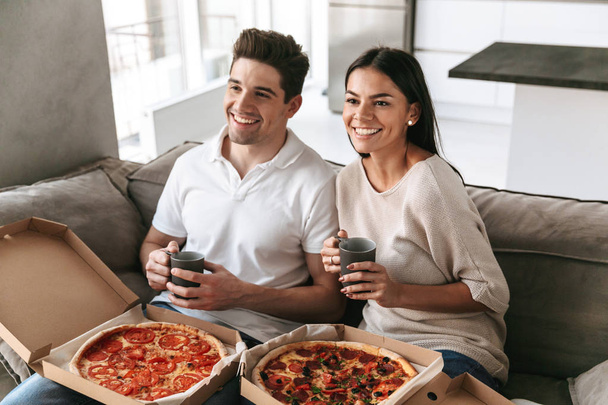 Cheerful young couple sitting on a couch at home, eating pizza, watching TV - Foto, afbeelding