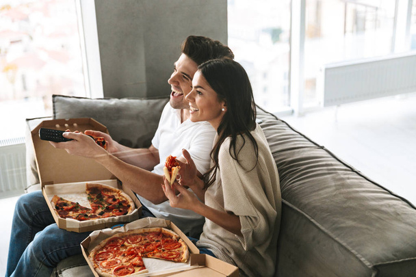 Cheerful young couple sitting on a couch at home, eating pizza, watching TV - Photo, image