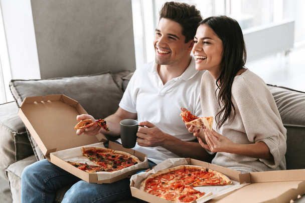 Cheerful young couple sitting on a couch at home, eating pizza, watching TV - Photo, Image