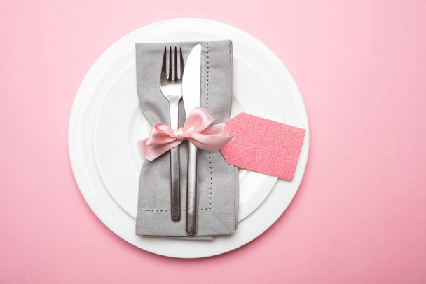 Table place setting in pink. Gray linen napkin with ribbon and blank tag on pink color background, top view, copy space - 写真・画像