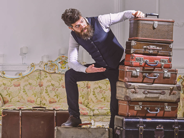 Man with beard and mustache packed luggage, white interior background. Move out, transportation concept. Macho elegant on shocked face, exhausted at end of packing, leans on pile of vintage suitcases - Фото, зображення