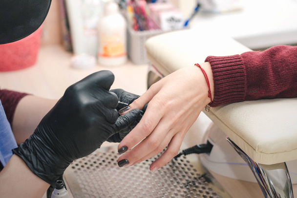 The manicurist in black latex gloves covers the client's nails with a colored gel polish in the beauty salon. - Fotografie, Obrázek