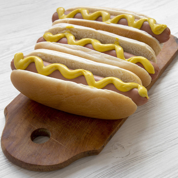 Hotdog mit gelbem Senf auf rustikalem Brett auf weißem Holztisch, Seitenansicht. Nahaufnahme. - Foto, Bild