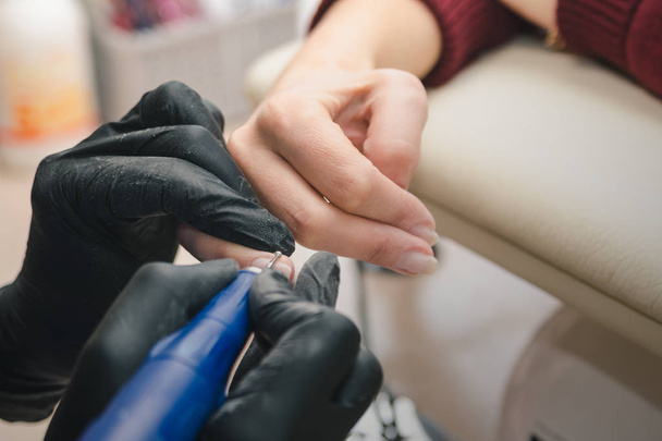 Een manicure in zwarte latex handschoenen maakt een hardware manicure naar een client met behulp van een handmatige elektrische freesmachine in een schoonheidssalon, close-up. - Foto, afbeelding