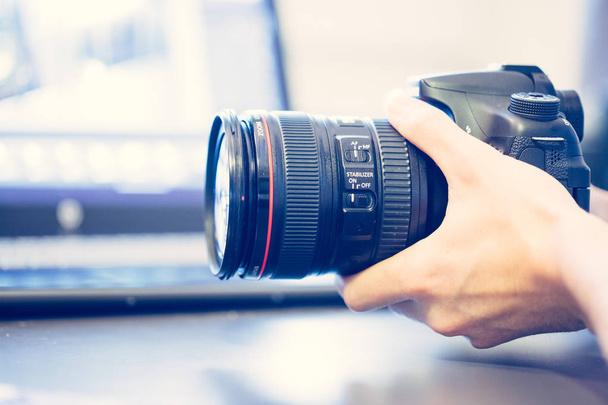 Photographer is holding a professional camera with telephoto lens in his hand, laptop in the blurry background - Foto, Bild