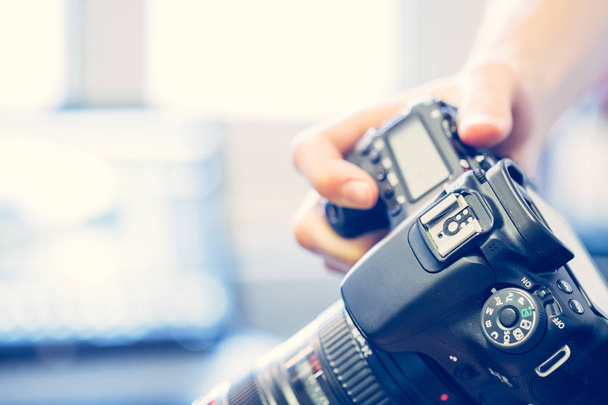 Photographer holds a reflex camera with telephoto lens in his hand. Table and laptop in the blurry background - Foto, imagen