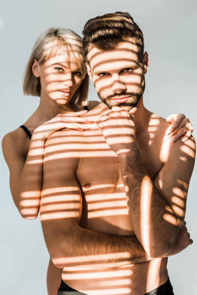 νεαρή γυναίκα αγκαλιάζει άνδρα με αυτοπεποίθηση shirtless απομονώνονται σε γκρι με σκιές - Φωτογραφία, εικόνα