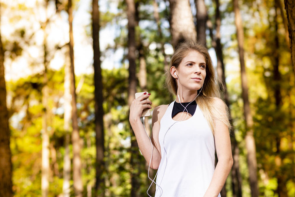 Linda modelo loira em roupas de ginástica ouvindo música em seu telefone celular. Floresta turva ao pôr-do-sol
 - Foto, Imagem