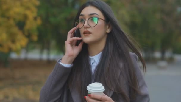 stylish businesswoman in modern coat and talks on cellphone walking in park - Felvétel, videó