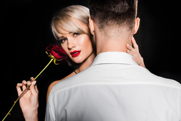 sensual girlfriend holding red rose flower while hugging boyfriend, isolated on black - Фото, изображение