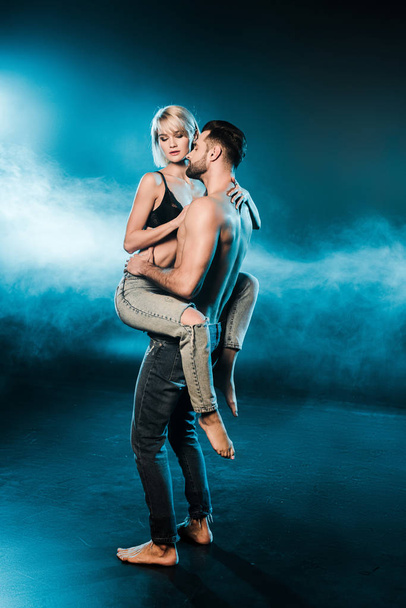 handsome seductive man holding blonde girlfriend on blue smoky background - Foto, imagen