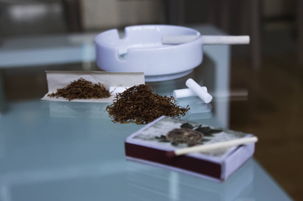 Un mazzo di tabacco marrone arrotolato a mano con fiammiferi e la sigaretta nel posacenere bianco sul tavolo di vetro Filtro per cesti di carta
 - Foto, immagini