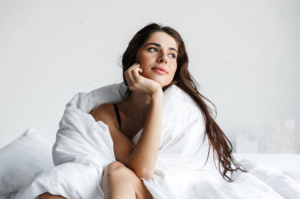 Mooie jonge vrouw die het dragen van zwarte lingerie zittend op bed bedekt met deken - Foto, afbeelding