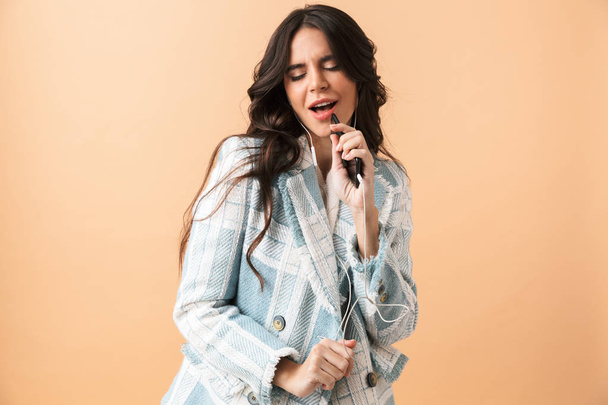 Beautiful brunette woman dressed in plaid jacket standing isolated over beige background, holding mobile phone, singing with earphones - Photo, image