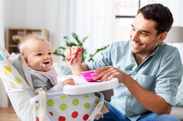 father feeding happy baby in highchair at home - Foto, Imagen