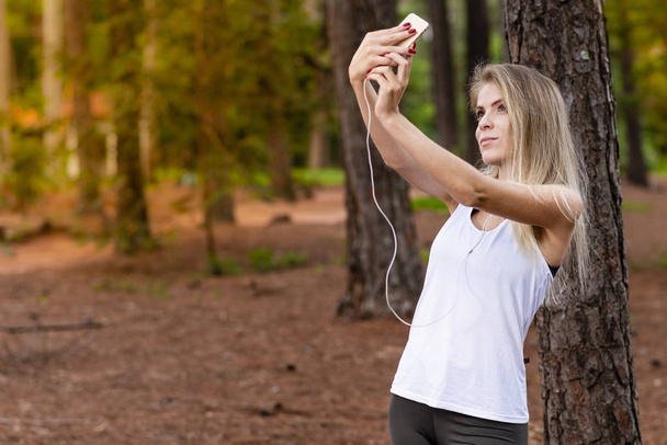 Hermoso modelo rubio en el bosque utilizando el teléfono celular con auriculares tirando selfie. Escuchando música
 - Foto, Imagen