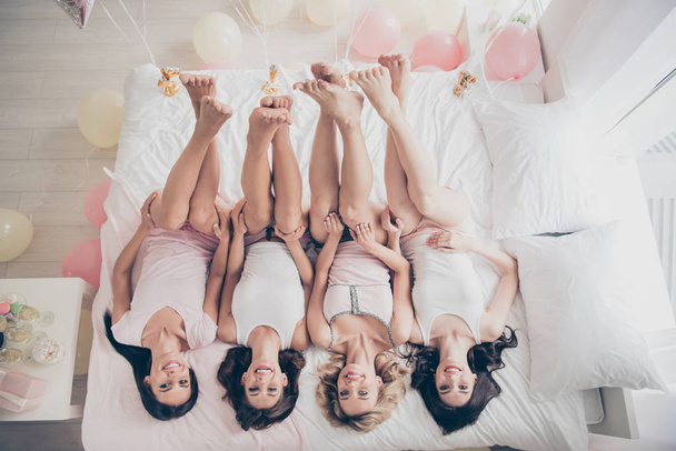 Top above high angle view of nice lovely attractive charming cheerful slim thin fit trendy girlfriends having fun lying on bed rising legs up in light white interior decorated house indoors - Фото, изображение