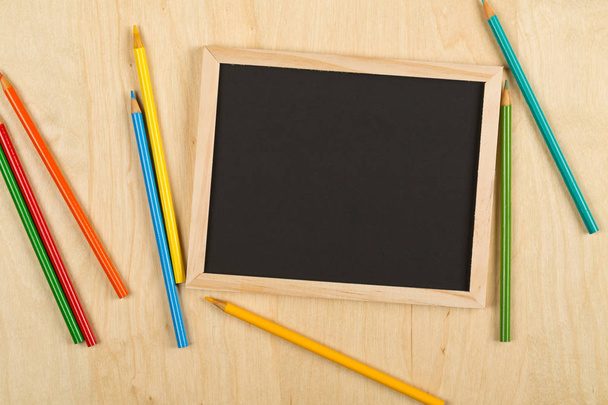Black, empty, blank chalkboard with colored pencils on brown wooden desk flat lay from above with copy space - Photo, image
