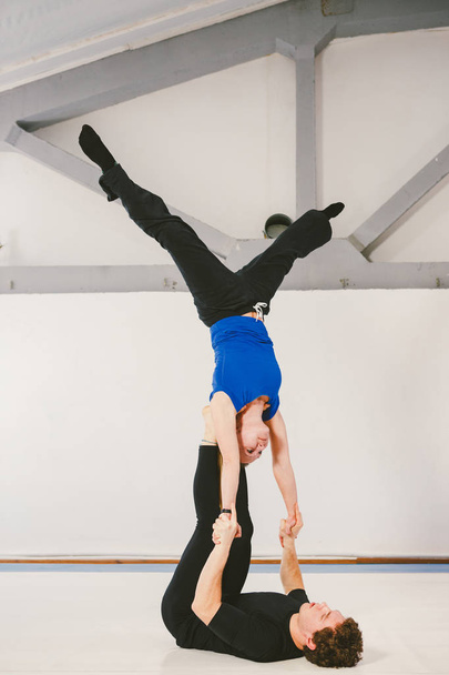 Theme is sports and acreage. A young Caucasian male and female couple practicing acrobatic yoga in a white gym on mats. a man lies on his back and holds a woman on extended arms. Pose HAND-TO-HAND - Foto, Imagen