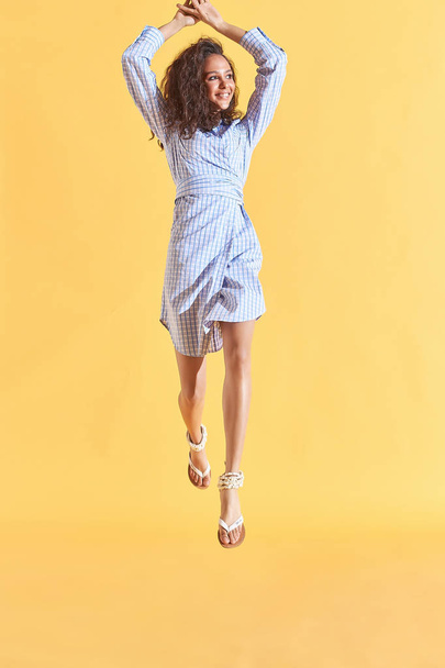 Full length portrait of tanned female model with curly brown hair, dressed in blue plaid shirt dress. Dynamic poses. Yellow background - Fotoğraf, Görsel