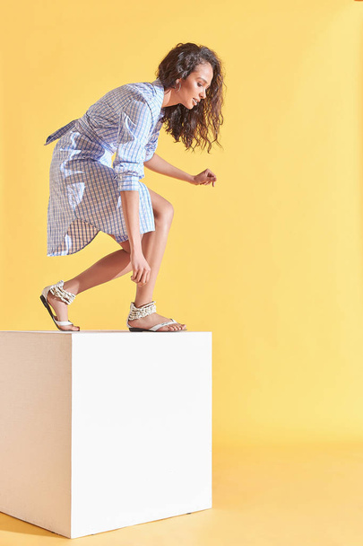 Fashion portrait of tanned female model with curly brown hair, dressed in blue plaid shirt dress jumping on white cube. Studio, yellow background - Zdjęcie, obraz