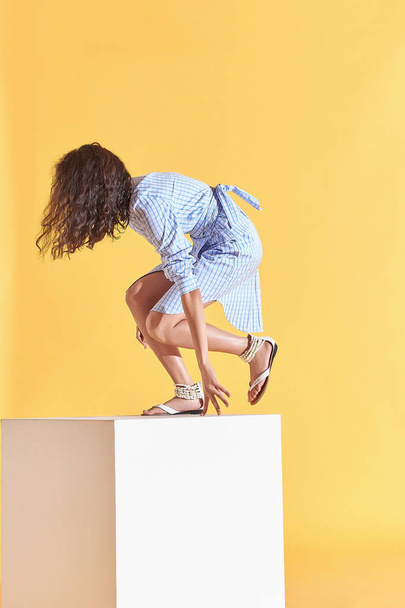 Fashion portrait of tanned female model with curly brown hair, dressed in blue plaid shirt dress jumping on white cube. Studio, yellow background - Foto, Imagen