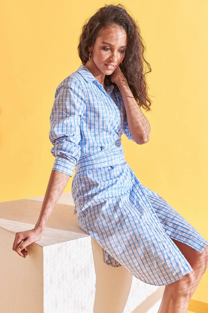 Fashion portrait of tanned female model with curly brown hair, dressed in blue plaid shirt dress sitting on white cube. Studio, yellow background - Fotó, kép