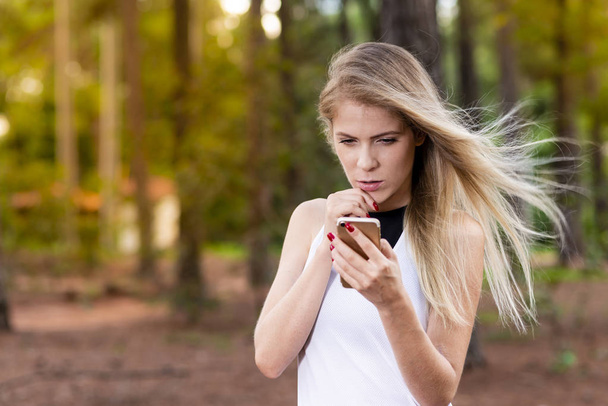 Beautiful blond young woman in park at sunset checking messages on her cell phone - Image
. - Фото, изображение