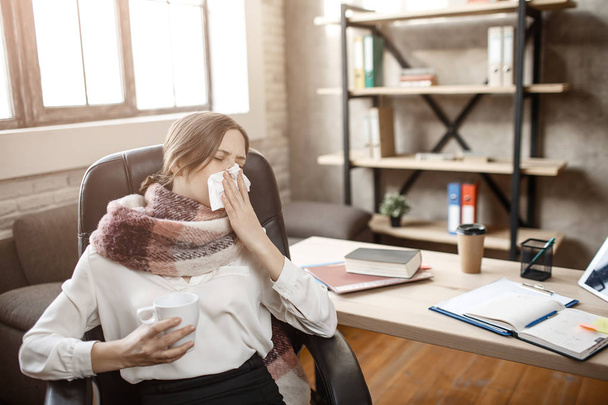Sick young businesswoman sit at table in room and sneezing. She suffer from disease. Woman cant work. - Photo, Image