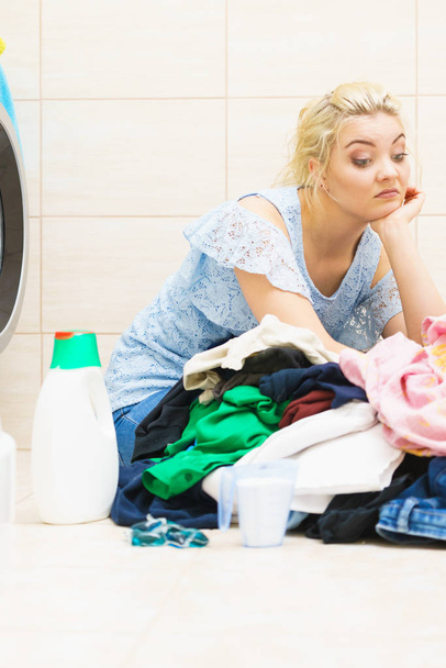 Sad woman having a lot of laundry to make. Unhappy female surrounded by big clothing piles stack sitting next to washing machine being tired houseworking. - 写真・画像