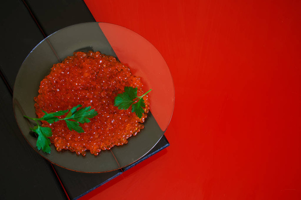 Red salmon caviar and parsley leaves in a transparent plate on a black and red background. Healthy food.                                - Photo, Image