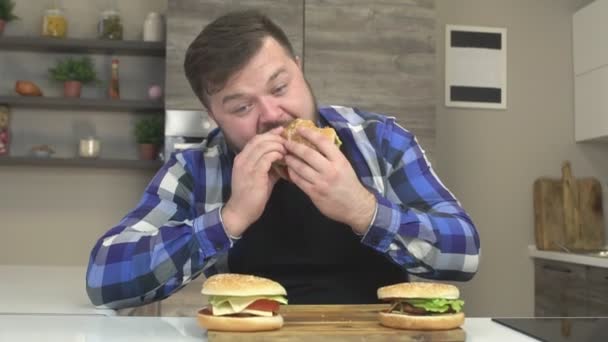 Fat man with a beard gorges on the kitchen fast food hamburgers, extra calories, slow motion, gluttony - Footage, Video