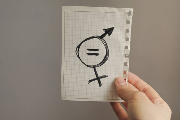 крупним планом молода біла жінка на вулиці, що показує аркуш паперу з символом гендерної рівності намальованої в ньому
 - Фото, зображення