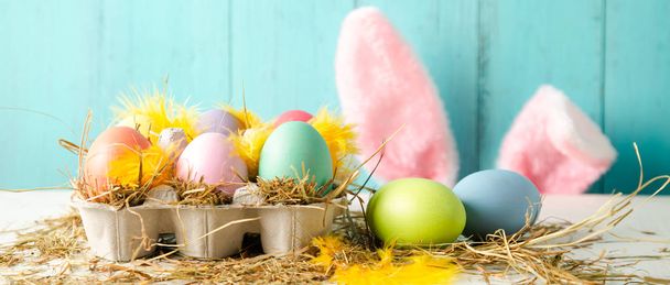 Pastel colored easter eggs in a egg box with hay and feathers as a nest and rabbit ears behind against a wooden turquoise background. Banner size - Photo, Image