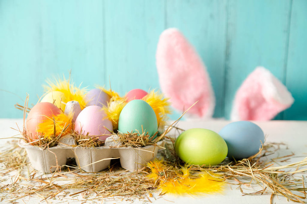 Pastel colored easter eggs in a egg box with hay and feathers as a nest and rabbit ears behind against a wooden turquoise background - Photo, Image