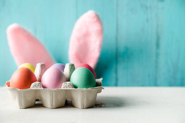 Pastel colored easter eggs in a egg box and rabbit ears behind with a wooden turquoise background - Photo, Image