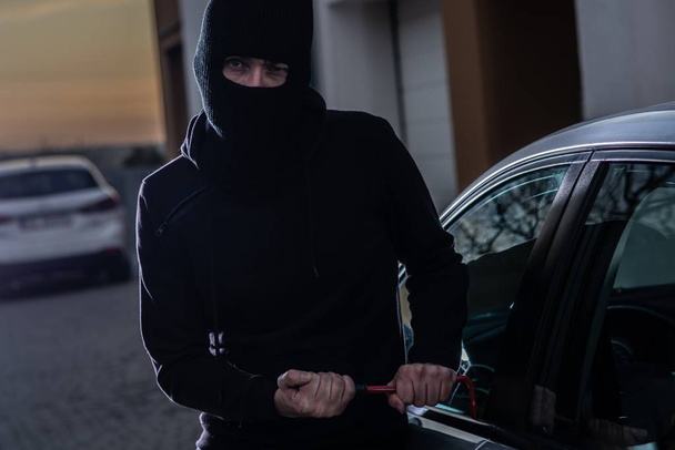 Car thief in a balaclava is trying to break into a car using a crowbar - Photo, Image