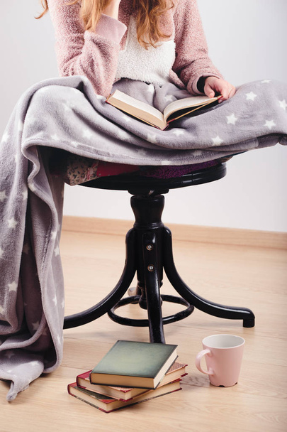 Girl enjoying the reading a book and drinking coffee at home. Young woman sitting on a chair, wrapped in blanket, holding book, relaxing at home. Portrait orientation. Front view - Photo, Image