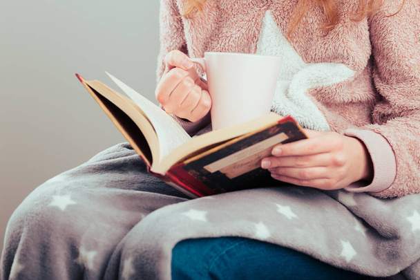 Girl enjoying the reading a book and drinking coffee at home. Young woman sitting on a chair, wrapped in blanket, holding book, relaxing at home. Portrait orientation. Front view - Zdjęcie, obraz
