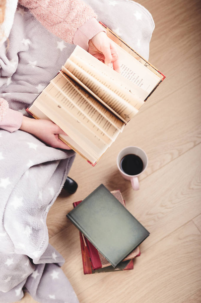 Girl enjoying the reading a book and drinking coffee at home. Young woman sitting on a chair, wrapped in blanket, holding book, relaxing at home. Portrait orientation. View from above - Foto, immagini