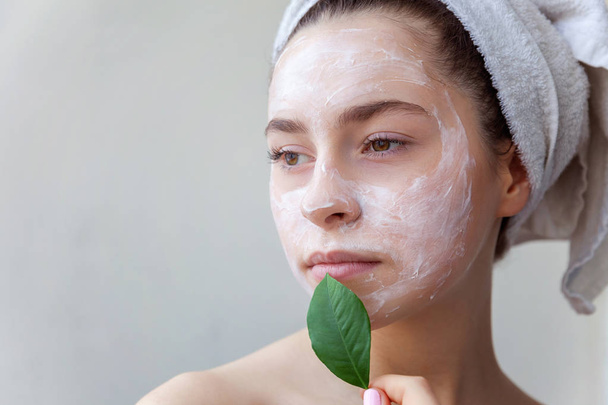 Beauty portrait of woman in towel on head with white nourishing mask or creme on face and green leaf in hand on white background isolated. Skincare cleansing eco organic cosmetic spa relax concept - Фото, изображение